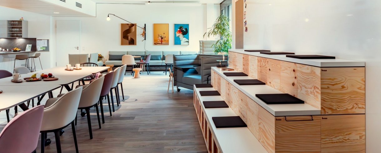 Im Vordergrund Sitztreppe und lange Tafel mit Stühlen. Im Hintergrund Gemälde und Aufenthaltsbereich.