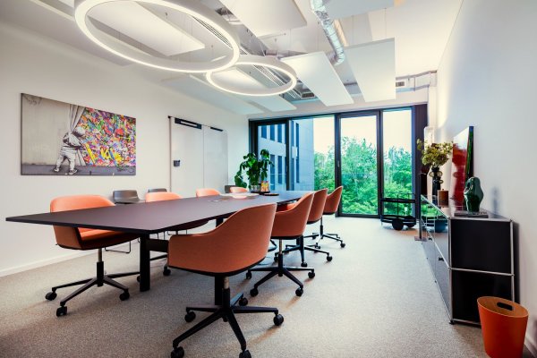 Ringlicht decke lange Tafel orangene Sessel mit Rollen Gemälde an Wand durchgehendes helles Fenster